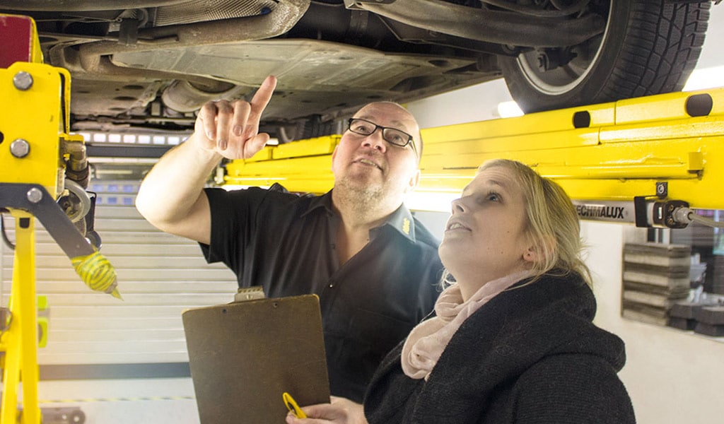 Zwei Personen stehen unter Hebebühne und begutachten ein Auto von unten.