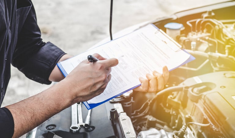 Person mit Klemmbrett und Checkliste über geöffneter Motorhaube.