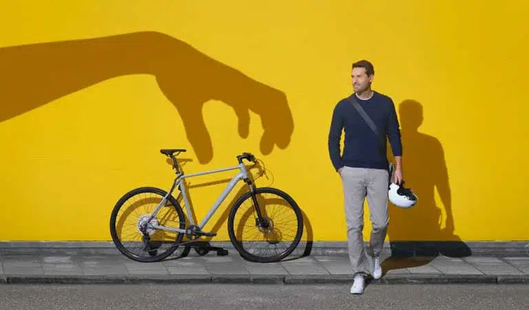 Ein Mann lächelnd, hält Helm neben Fahrrad, gelbe Wand, Schatten einer großen Hand.