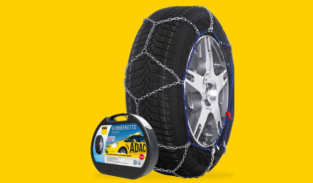 Reifen mit Schneeketten auf gelbem Hintergrund, silberne Felge, schwarze Reifen, Verpackung daneben.