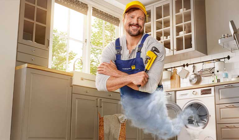 Mann in blauem Overall, lächelt und schwebt als Gini aus Waschmaschine, hält Rohrzange, helle Küche.