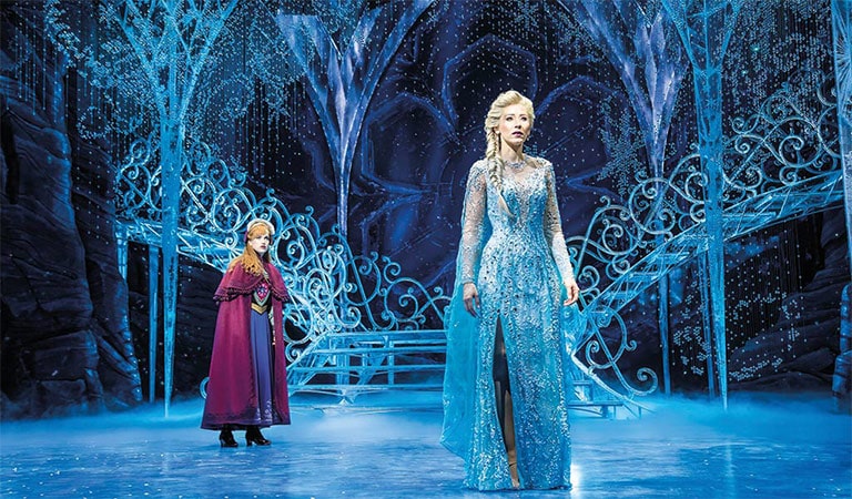 Zwei Figuren in märchenhafter Eislandschaft; vorne blond, blaues Kleid; hinten rotes Kleid, staunend.