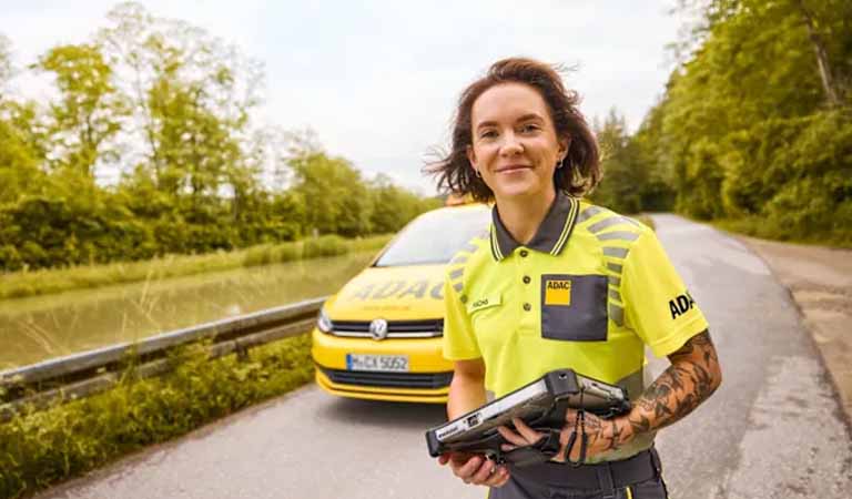 Eine lächelnde Frau in ADAC Uniform vor einem gelben Auto auf einer Landstraße.