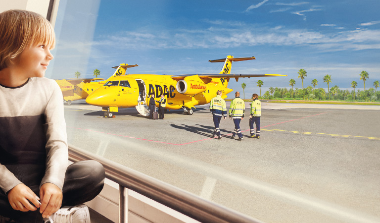 Ein lächelndes Kind blickt auf ein gelbes ADAC Flugzeug mit Personal auf dem sonnigen Rollfeld.