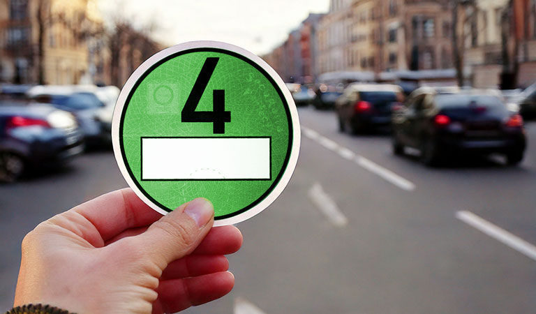 Hand hält grüne Umweltplakette vor belebter Straße in Stadtumgebung.