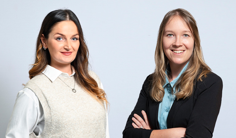 Zwei lächelnde Frauen, die in die Kamera schauen, eine mit langem Haar und Pullover, die andere im Anzug.