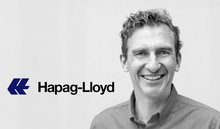 Ein lächelnder Mann mit lockigem Haar trägt ein Hemd vor dem Hapag Lloyd Logo