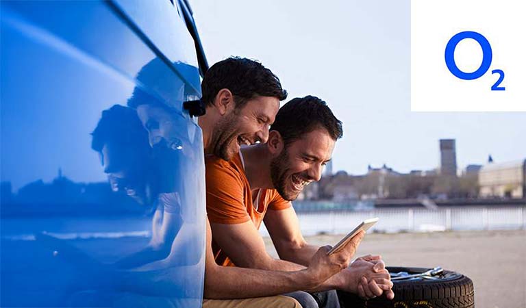 Zwei lachende Männer sitzen neben einem blauen Auto und schauen fröhlich auf ein Smartphone