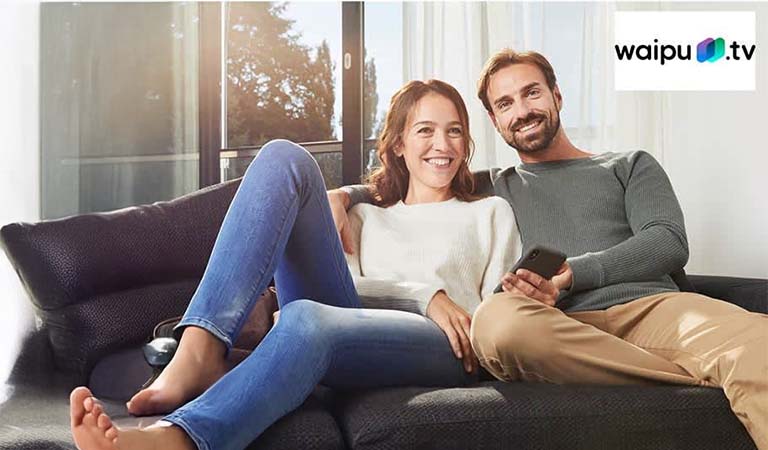 Ein lächelnder Mann und eine Frau sitzen entspannt auf einem dunklen Sofa im Sonnenlicht