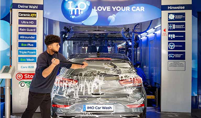 Ein junger Mann mit lockigem Haar wäscht ein silbernes Auto mit viel Schaum