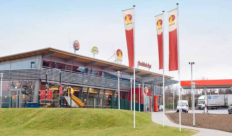 Eine moderne Raststätte mit roten Elementen, Fahnen und einem Spielplatz draußen