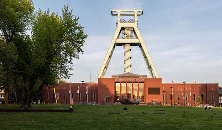 Das rote Ziegelgebäude mit großem Förderturm ragt vor grünem Rasen und Bäumen heraus