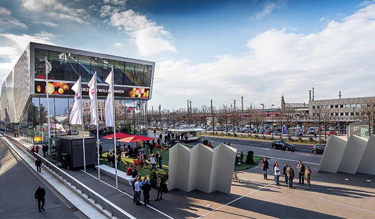 Eine Gruppe von Menschen versammelt sich vor einem modernen Glasgebäude mit freundlicher Stimmung und mehreren weißen Fahnen bei sonnigem Wetter
