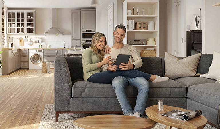 Ein lächelndes Paar sitzt entspannt auf einem grauen Sofa und betrachtet gemeinsam ein Tablet