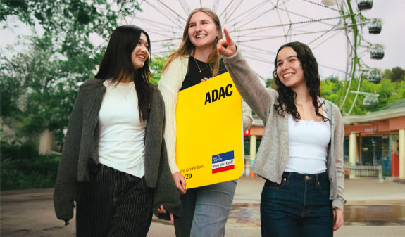 Drei junge Frauen lächeln fröhlich im Freizeitpark mit großer gelber ADAC Mitgliedskarte