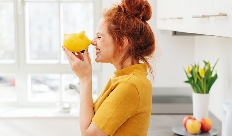 Eine lachende Frau mit roten Haaren hält ein gelbes Sparschwein in einer hellen Küche