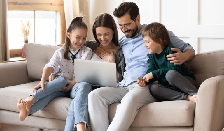 Eine lächelnde Familie mit zwei Kindern sitzt eng auf einem beigen Sofa vor einem Laptop