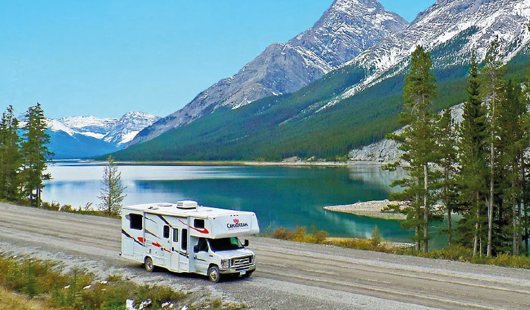 Ein weißes Wohnmobil fährt an einem klaren See mit Bergen und Tannen vorbei