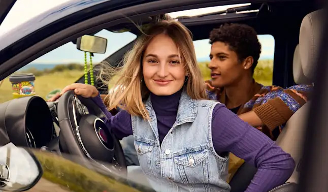 Junge Frau mit blondem Haar lächelt freundlich im offenen Auto, junger Mann daneben
