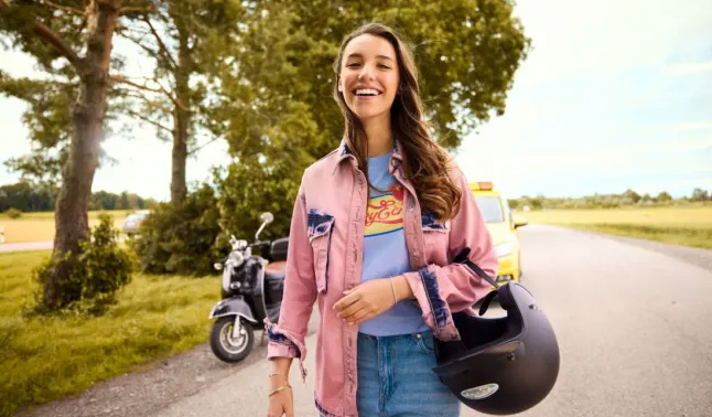 Junge Frau mit fröhlichem Gesichtsausdruck in Jeans und rosa Jacke hält Motorradhelm