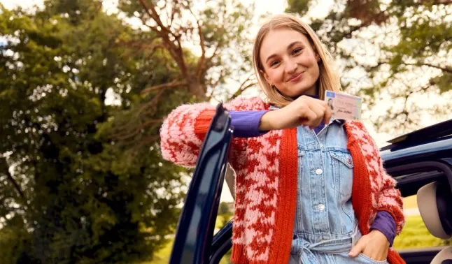 Junge Frau mit blonder Frisur lächelt stolz und hält einen Führerschein am Auto hoch