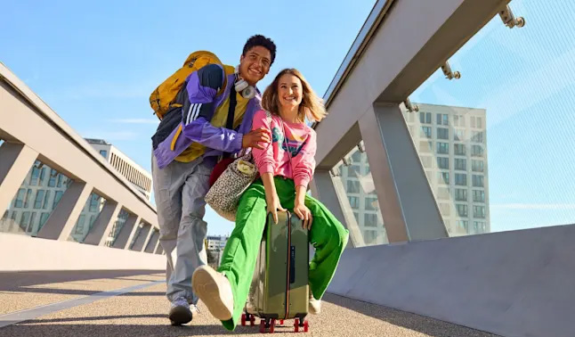 Zwei junge Erwachsene lächeln fröhlich auf einer Brücke mit buntem Gepäck