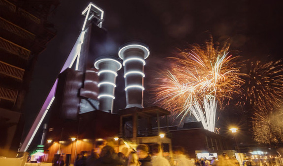 Nachts beleuchtetes Industriegebäude mit weißen Lichtern und buntem Feuerwerk