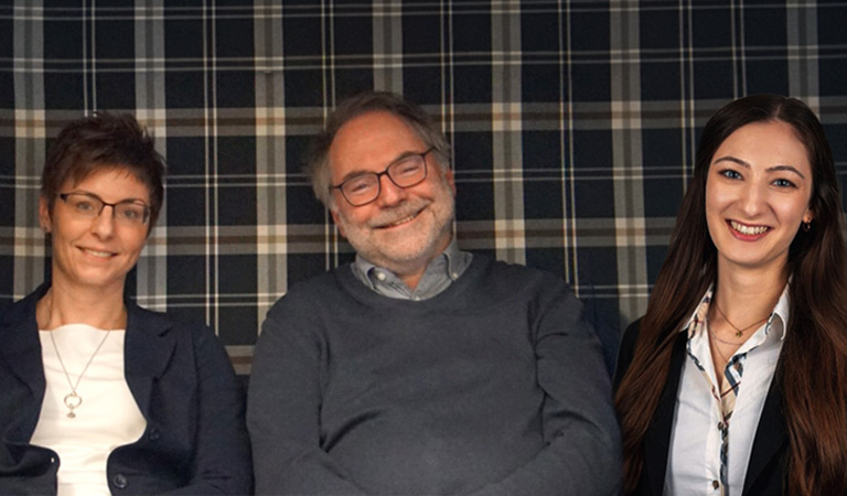 Zwei Mitarbeiterinnen und ein Mitarbeiter des ADAC Reisebüros Bochum lächeln vor einer Wand mit Karomuster: S. Himmerich, N. Martin und D. Schnegelsberg.