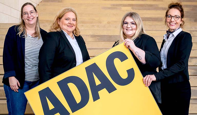 Vier Mitarbeiterinnen des ADAC Reisebüros Siegen in Businesskleidung halten ein großes gelbes ADAC Schild: A. Spielmann, H. Schröder, V. Müller und J. Pursch.
