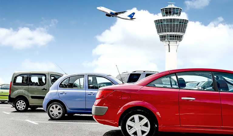 Mehrere bunte Autos stehen geparkt vor einem modernen Flughafentower, oben fliegt ein Flugzeug