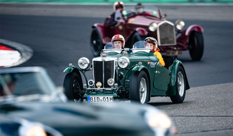 Zwei ältere Personen lächeln entspannt im grünen Oldtimer auf einer Rennstrecke