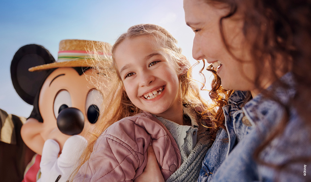 Eine junge Frau lacht fröhlich, während sie neben einem Kind mit lockigem Haar steht, im Hintergrund ist eine große Mickey Maus Figur mit Strohhut zu sehen