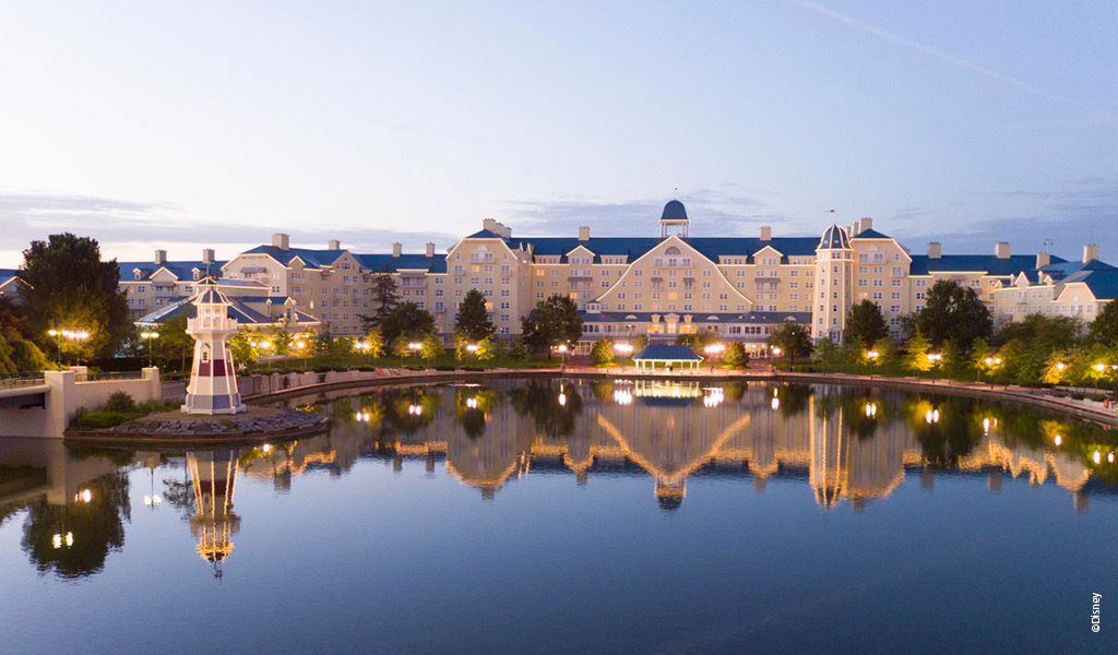 Ein großes, gelbes Hotel spiegelt sich im ruhigen See, Bäume und Lichter flankieren