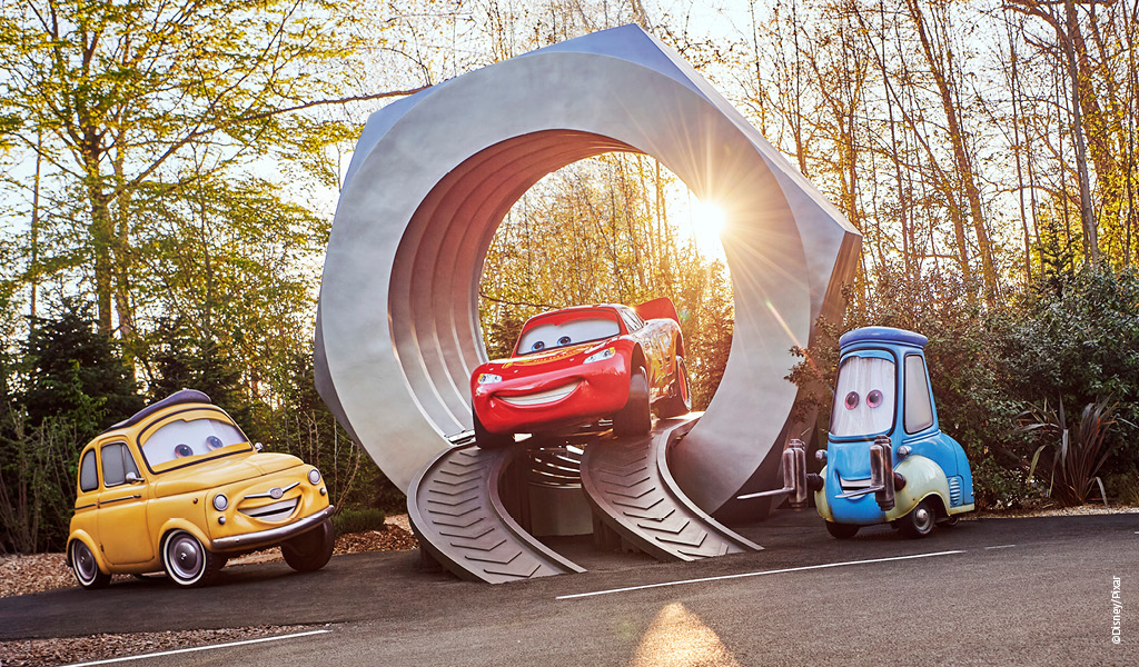 Drei animierte Autos mit fröhlichen Gesichtern vor einer großen silbernen Skulptur
