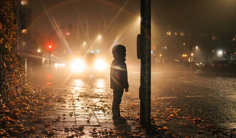 Ein Kind steht im Herbstnebel am Straßenrand mit Kapuze beleuchtet von Autoscheinwerfern
